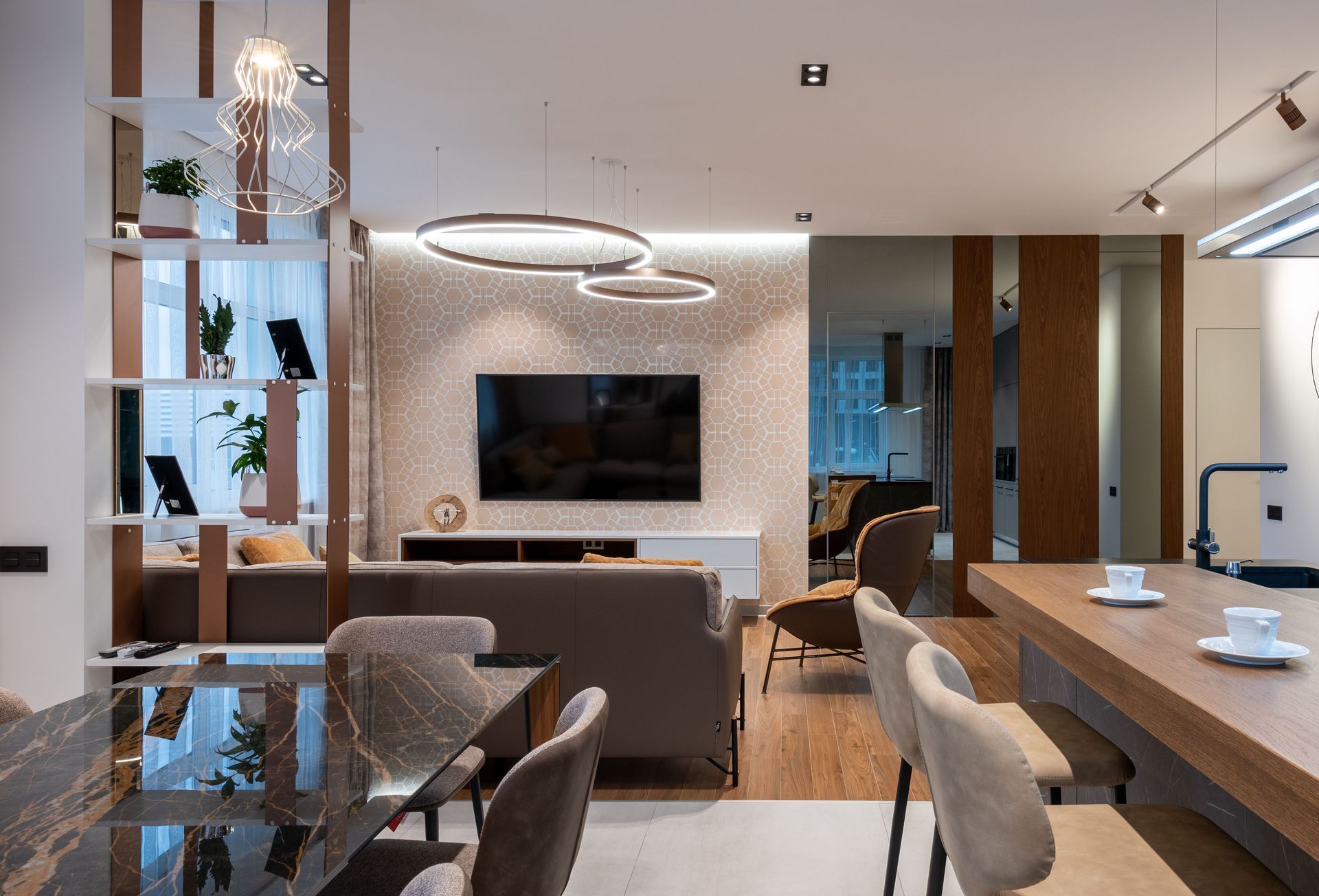 Room with couch and pillows and TV on wall under cabinet near armchair and mirror near window with curtains and shelves near counter with coffee cups and chairs near table